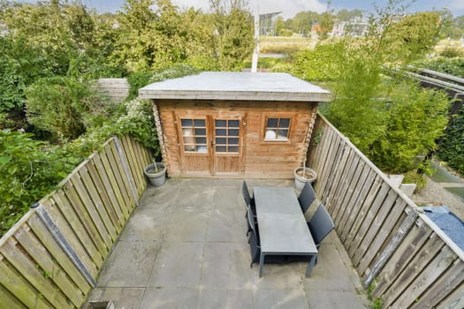 A stylish wooden garden shed surrounded by greenery, featuring a patio area with a table and chairs, perfect for outdoor relaxation and storage.
