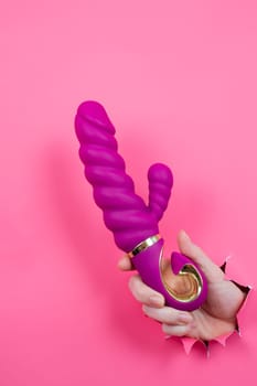 Female hand sticking out with pink sex toy from pink paper background. Copy space. Vertical photo