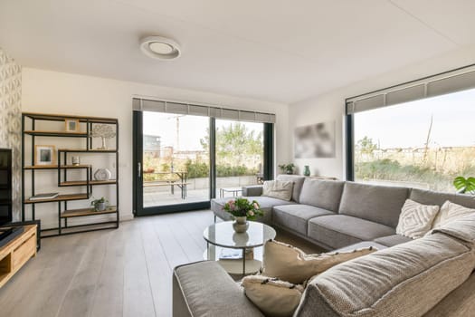 An elegant modern living room featuring minimalist furniture with a spacious layout, natural light, and greenery visible outside through large glass windows.