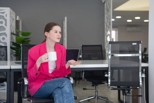 Caucasian business woman in fuchsia jacket drinking coffee at her desk