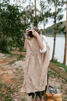 A young woman dressed in natural, earthy tones stands near a tranquil lake, emotionally engaged as she prepares to wear a dress, reflecting a love for nature and fashion