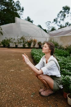 A young woman squatting in a lush garden, dressed in a casual white shirt and green shorts, exuding joy and connection to nature, emphasizing eco friendly living