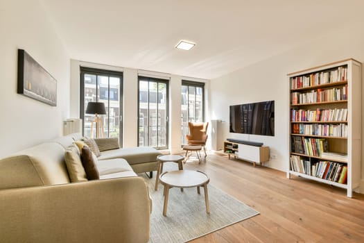 Bright and spacious modern living room featuring a cozy sofa, wooden flooring, a stylish bookshelf, and large windows for natural light.