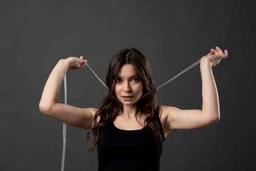 Young woman with slim skinny body wear black t-shirt hold measure tape on neck look in a camera isolated on dark grey background. Diet and weight loss