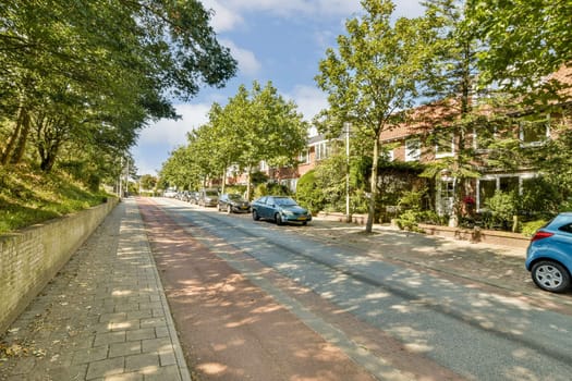A peaceful residential street lined with trees and vibrant houses. Bright blue skies peek over the neighborhood, creating a picturesque scene adorned with cars parked along the road.