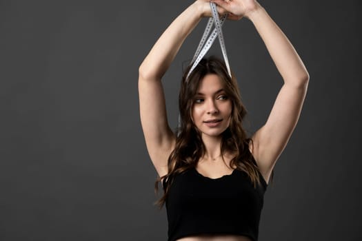 Young woman with slim skinny body wear black t-shirt hold measure tape across a neck isolated on dark grey background. Diet and weight loss