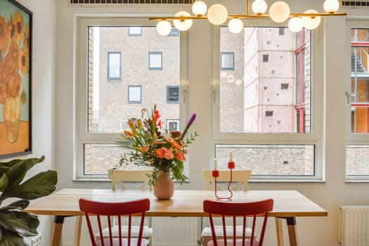 A stylish dining area featuring a wooden table adorned with a vibrant floral arrangement, surrounded by contemporary chairs and large windows.