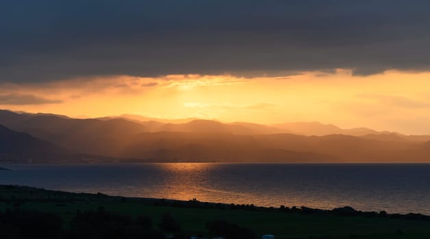 A breathtaking view unfolds as the sun sets beyond the distant mountains, casting golden rays across the calm water and enveloping the landscape in warm colors.