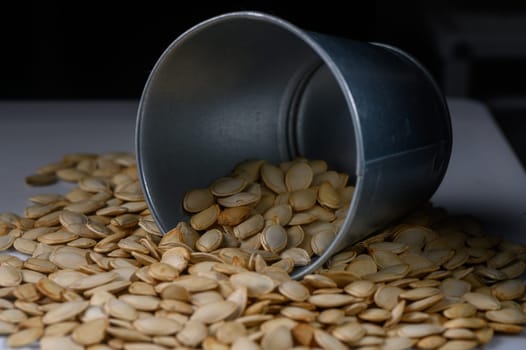 A silver bucket tips over, spilling a cascade of beige pumpkin seeds onto a wooden surface. The warm, earthy tones evoke a cozy autumn atmosphere perfect for harvest celebrations.