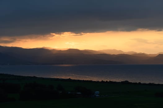 A breathtaking view unfolds as the sun sets beyond the mountains, casting warm golden light across the sea and illuminating the cloud-draped sky.