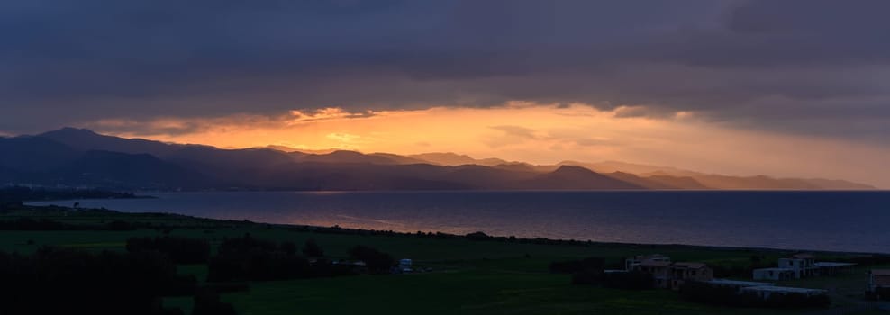 As daylight fades, a breathtaking array of colors emerges over the sea, illuminating the rugged mountains while casting soft shadows on the landscape. A moment of serene beauty.