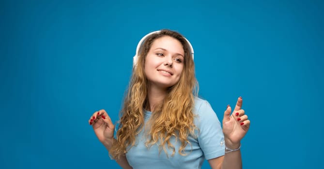Cheerful young woman wearing blue t-shirt listening to music with headphones, dancing isolated over blue background