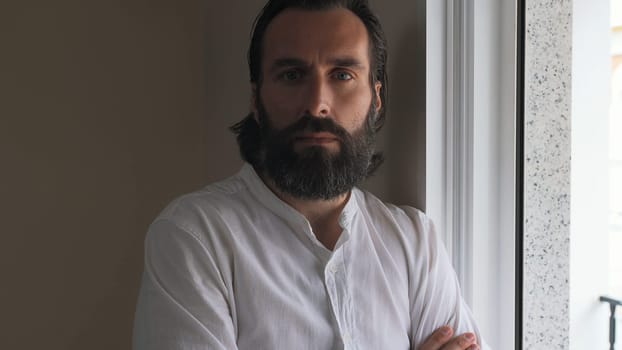 A confident and composed young man with a beard stands indoors, gazing thoughtfully.