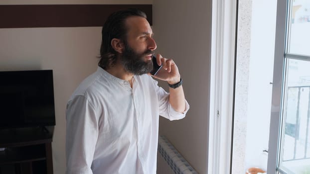 Young man standing beside a window, speaking on his smartphone with a focused expression. Wearing a white shirt, he appears businesslike in his home office setting.