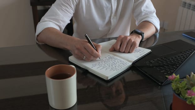 A person sits at a desk in a modern office environment, writing in a notebook. The workspace features a laptop, a coffee mug, and a neat arrangement of essentials, creating a productive scene.