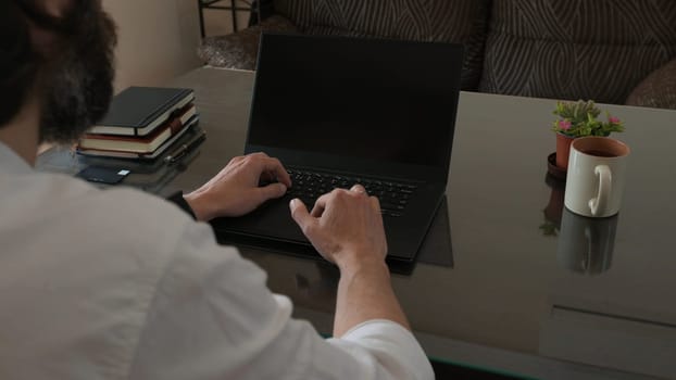 A man in a professional workspace uses a laptop computer while surrounded by notebooks, a plant, and a coffee mug. The scene suggests focus, productivity, and a comfortable office environment.