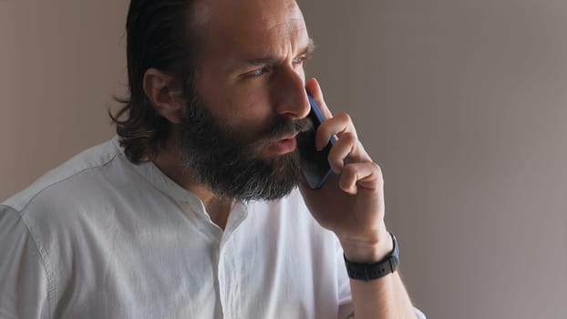 A focused man with a beard is using a smartphone, engaging in a conversation. Wearing a casual white shirt indoors, the scene captures a moment of concentration during communication.