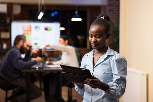 Professional black woman checks financial reports on her tablet while colleagues discuss business strategies. The modern office, late night setting and teamwork highlight productivity