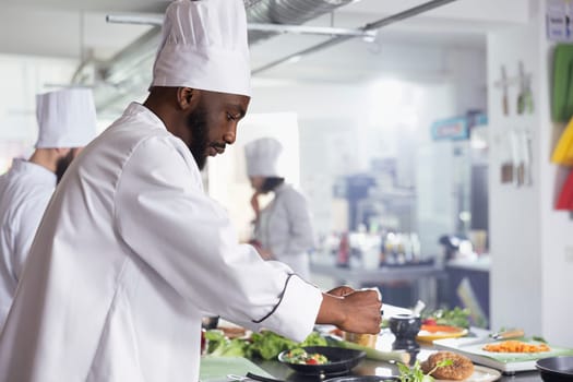 African american chef cooking delicious meals with rosemary or basil in a restaurant kitchen, sprinkles herbs leaves on the stove. Young cook using fresh produce and raw materials.