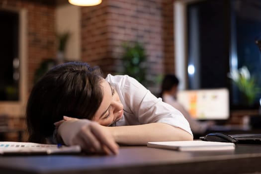 Tired businesswoman takes powernap at her workstation, resting briefly amid her demanding schedule in late night office setting. Caucasian entrepreneur dozes off at desk, exhausted from her workload.