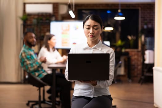 Focused asian businesswoman working on laptop, analyzing marketing trends as her coworkers discuss financial charts in background. The late night office setting highlights productivity.
