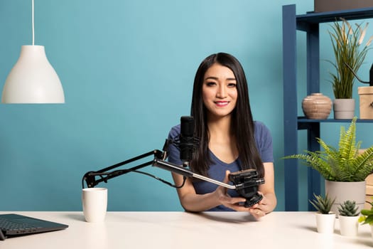 Asian female vlogger presenting a camera tripod as she speaks into a microphone in a professional studio setup. Young woman seated at her desk, reviewing filming equipment for content creation.