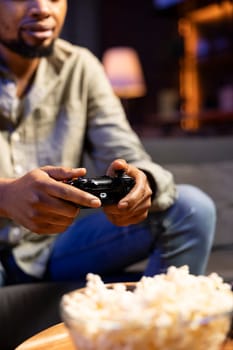 Black player enjoying shooter competition with popcorn at home, participating in online video games tournament with friends during game night. Man having fun with championship. Close up.