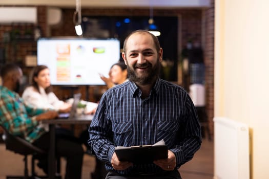 Caucasian male entrepreneur holds a clipboard in modern startup office. The night shift setting, brick wall and blurred team in background emphasize productivity and teamwork.