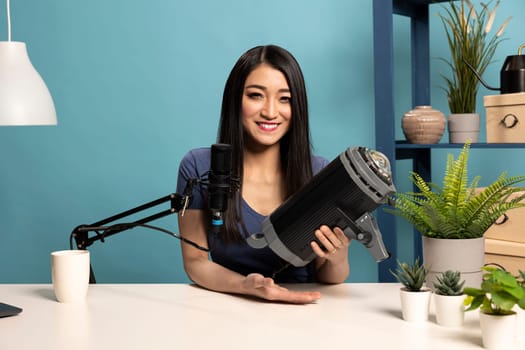 Cheerful female blogger holds up a strobe light in her studio, reviewing its features as she records social media content. She sits at desk with a microphone, engaging with her online audience.