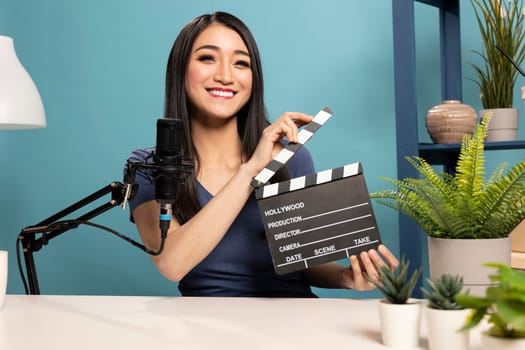 Female vlogger presents a clapperboard into the camera, sitting at a desk with a microphone. The scene highlights filmmaking equipment and atmosphere of video content creation for online reviews.