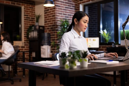 Professional woman working late at night in a modern office, seated at desk, focused on her laptop. Asian female entrepreneur using her personal computer, checking emails and managing business tasks.