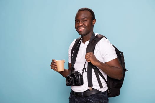 Smiling male model with a backpack and disposable cup looking directly at the camera, exuding confidence and readiness for a new creative project. Black photographer standing with his gear and coffee.