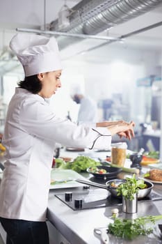 Young woman chef in restaurant kitchen carefully sprinkles fresh herbs onto a dish, enhancing the flavors with parsley, rosemary and basil. Modern workspace filled with fresh produce and spices.