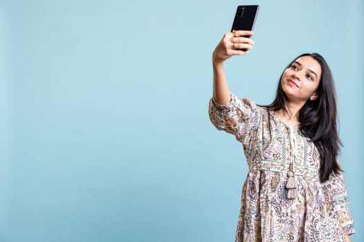 Jolly indian woman smiling for selfies posting them on social media profile, feeling confident and pretty in the studio. Happy silly girl taking pictures using phone selfie camera, blue background.