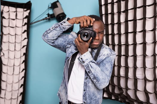 African American cameraman using his DSLR, highlighting his photography expertise. Young black photographer stands in studio, equipped with his camera gear and ready for photoshoot session with client.