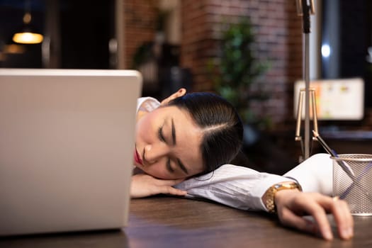 Asian female entrepreneur falls asleep at her desk during a late night shift in brick wall office. Tired businesswoman takes a brief rest amidst her busy schedule as her laptop remains open on table.