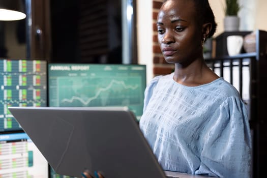 Female professional analyzes stock market movements on multiple screens, recording insights on laptop, engaging in trading session. Closeup on black woman focused on personal computer in modern office.