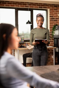 African american woman in workspace, looking excited and prepared for her role at creative agency. Portrait of black manager standing in brick wall office, holding clipboard and smiling at camera.