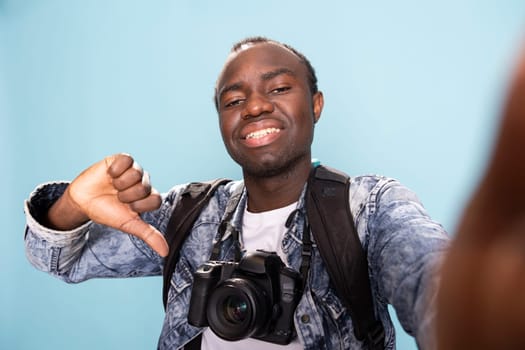 Happy african american photographer with a professional camera around his neck, giving thumbs down in studio. POV of a black male vlogger recording himself while making a playful disagreeing gesture.