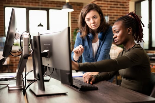 Caucasian and african american female colleagues working together at desk, refining digital art on desktop pc. Black employee touching computer screen during discussion with white woman manager.