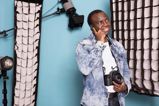 African american man grins widely while speaking on his mobile, holding his dslr device with one hand. Joyful male photographer with a camera around his neck stands, having a phone call in studio.