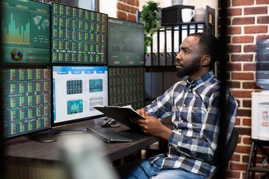 Male stock market trader with a clipboard in hand, analyzing real time data on a multi monitor setup in a dimly lit office. The scene reflects financial analysis and late night investment decisions.