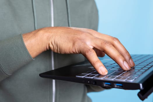 African american young guy typing on his laptop against blue background, composing emails and working on paperwork. Male overjoyed person multitasking in studio, cheerful adult. Close up.