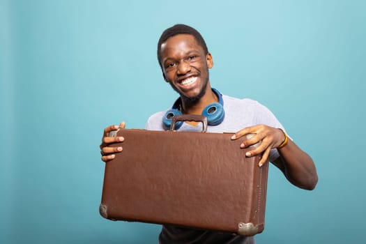 Cheerful african american man with a suitcase and headphones, excited for his journey. Black male tourist looking at the camera and posing with his luggage against an isolated background.