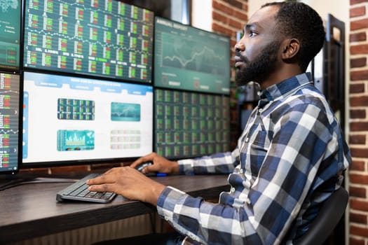 Focused male employee closely monitoring company shares, tracking growth on multiple screens. Black man sitting at his desk, deeply engaged in forex trading in a tech savvy workplace.