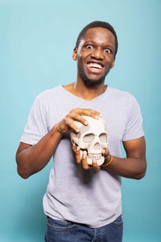 African american man with wide eyes, holding a skull and making goofy expressions as he looks at camera. Young black male individual grasping a human skeletal head against an isolated blue background.