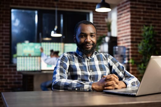 Portrait of african american man smiling and looking at the camera, seated at a desk with a laptop. The office is dimly lit with a brick wall, ideal for night shift work and stock market monitoring.