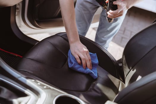 Portrait of one young Caucasian unrecognizable man who is bending over, polishing the front passenger leather seat inside the car with a blue rag on a sunny spring day at a car wash, side view close-up. Concept car wash, working people.