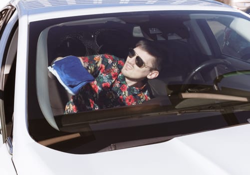 Portrait of one young Caucasian handsome man in sunglasses who washes windshield with blue cloth sitting inside car on sunny spring day at car wash, side view close-up. Concept car wash, working people.