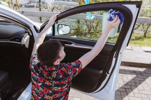 Portrait of a young Caucasian man sitting from the back and washing the glass from the inside of the car door with a blue rag on a sunny spring day at a car wash, side view close-up. Concept time car wash, working people.